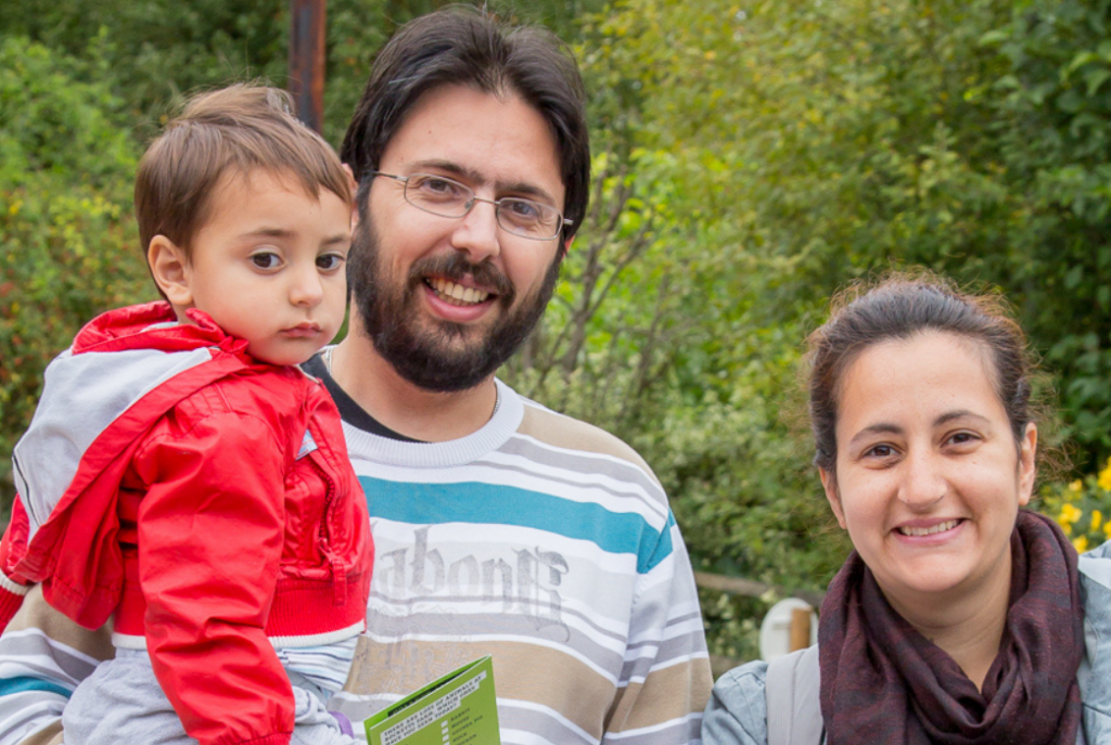 A woman has her hands resting on a pushchair and stands next to a man, who is holding a child in his arms. The man and woman are smiling at the camera, with green trees and bushes out of focus behind them. At the bottom of the photo is a semi-translucent pink banner, with white text reading: "CLAPA Coffee Club: 'Celebrating Difference'. Monday 10th May at 7pm on Zoom." The Awareness Week logo is in the bottom right and white CLAPA logo is in the top right of the photo