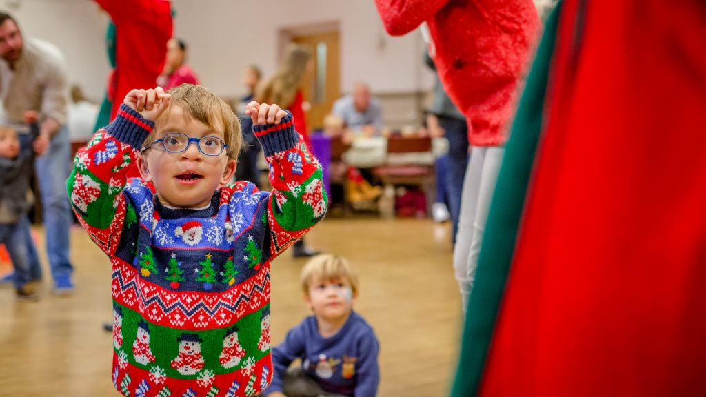 A young boy wearing glasses has his arms raised by his head and is staring up at someone. He is wearing a Christmas jumper