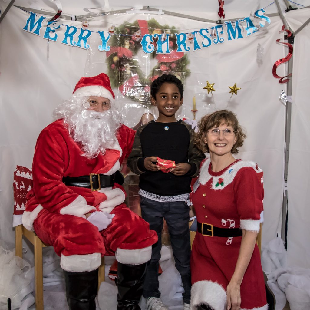 A boy is standing in front of a sign saying 'Merry Christmas' and is holding a small red present, next to a man dressed in a Santa costume and a woman dressed up in a red dress with a white trim.