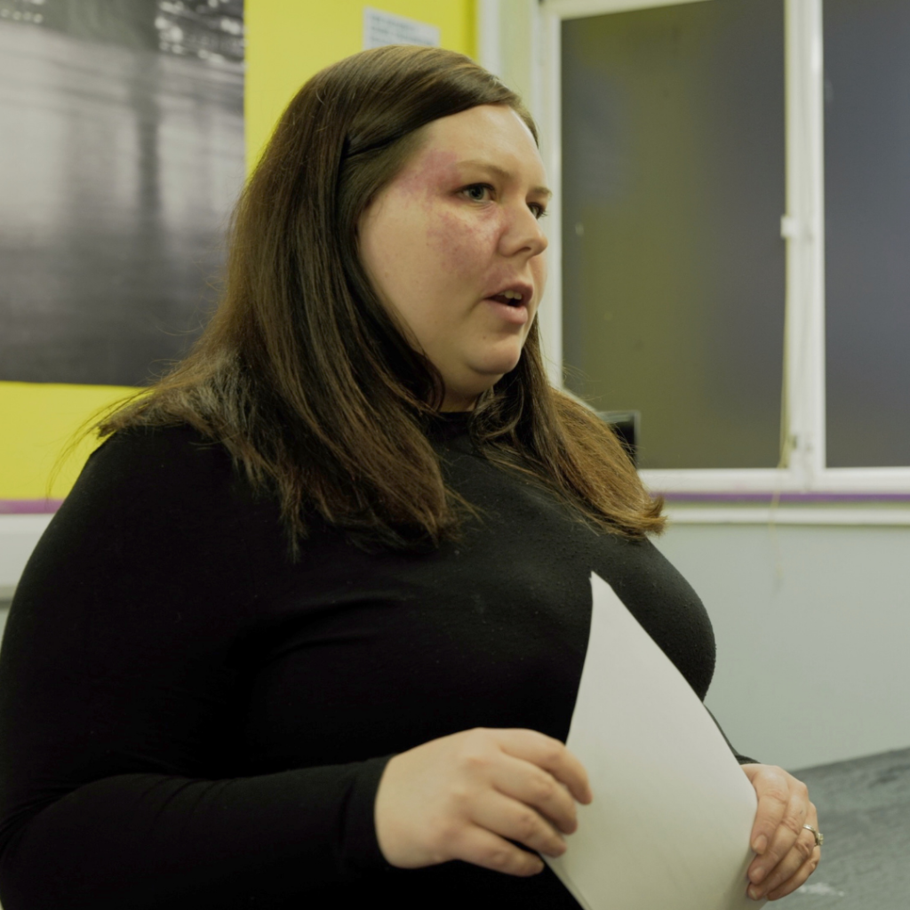 [Image ID: A female youth worker wearing a black jumper, holding a piece of white paper in a classroom setting.]