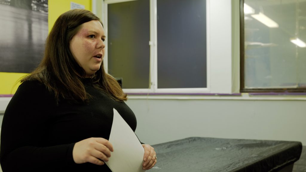 [Image ID: A female youth worker wearing a black jumper, holding a piece of white paper in a classroom setting.]