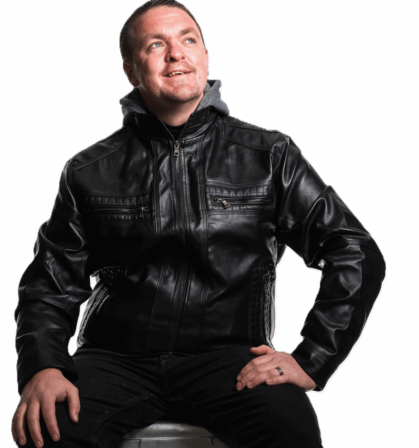 A man wears a leather jacket and is sat on a stool. He is looking up and smiling. He has a repaired cleft