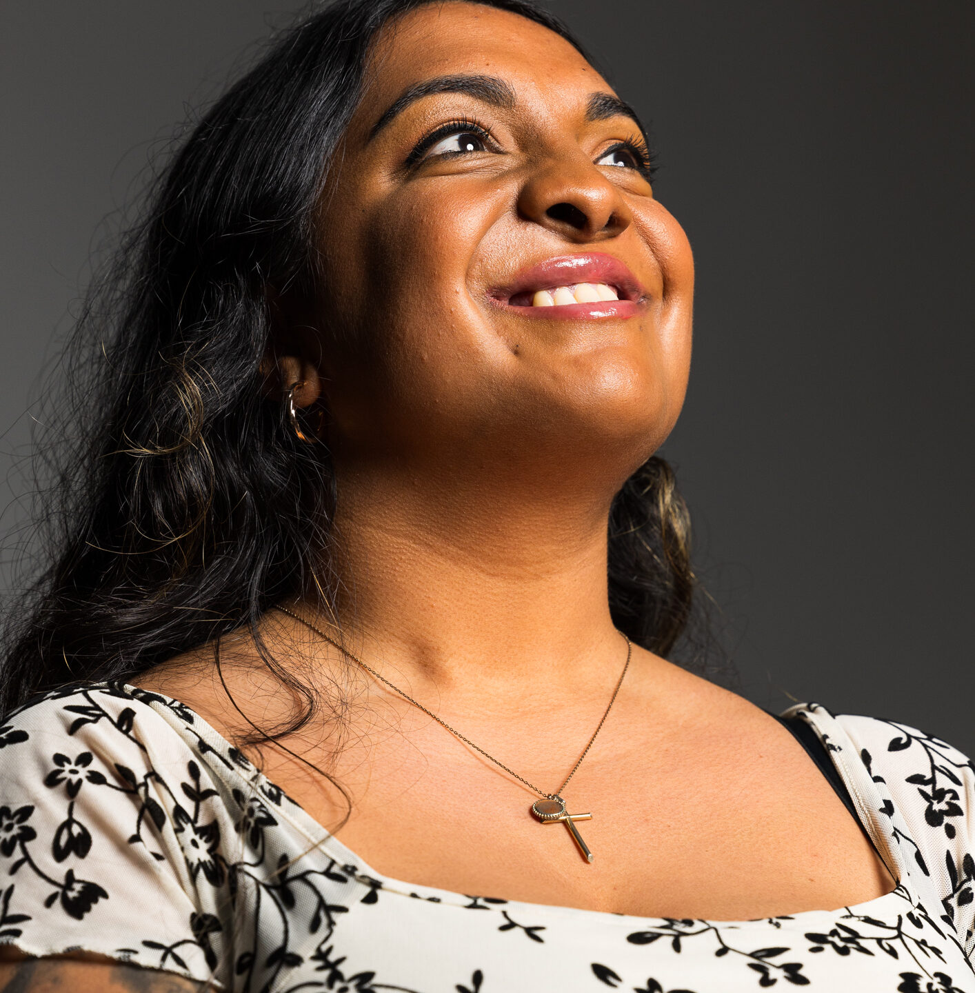 A lady of south asian heritage wears a black and white flowery dress. She is looking up off camera and smiling. She has a repaired cleft.