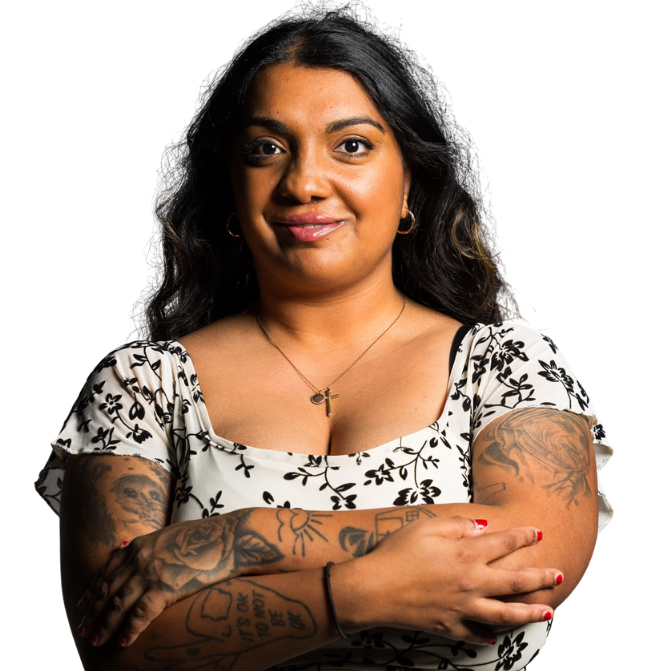 An adult lady with a cleft of Indian origin has her arms folded. She has tattoos on her arms and looks to the camera with a wry smile. She has long black hair.